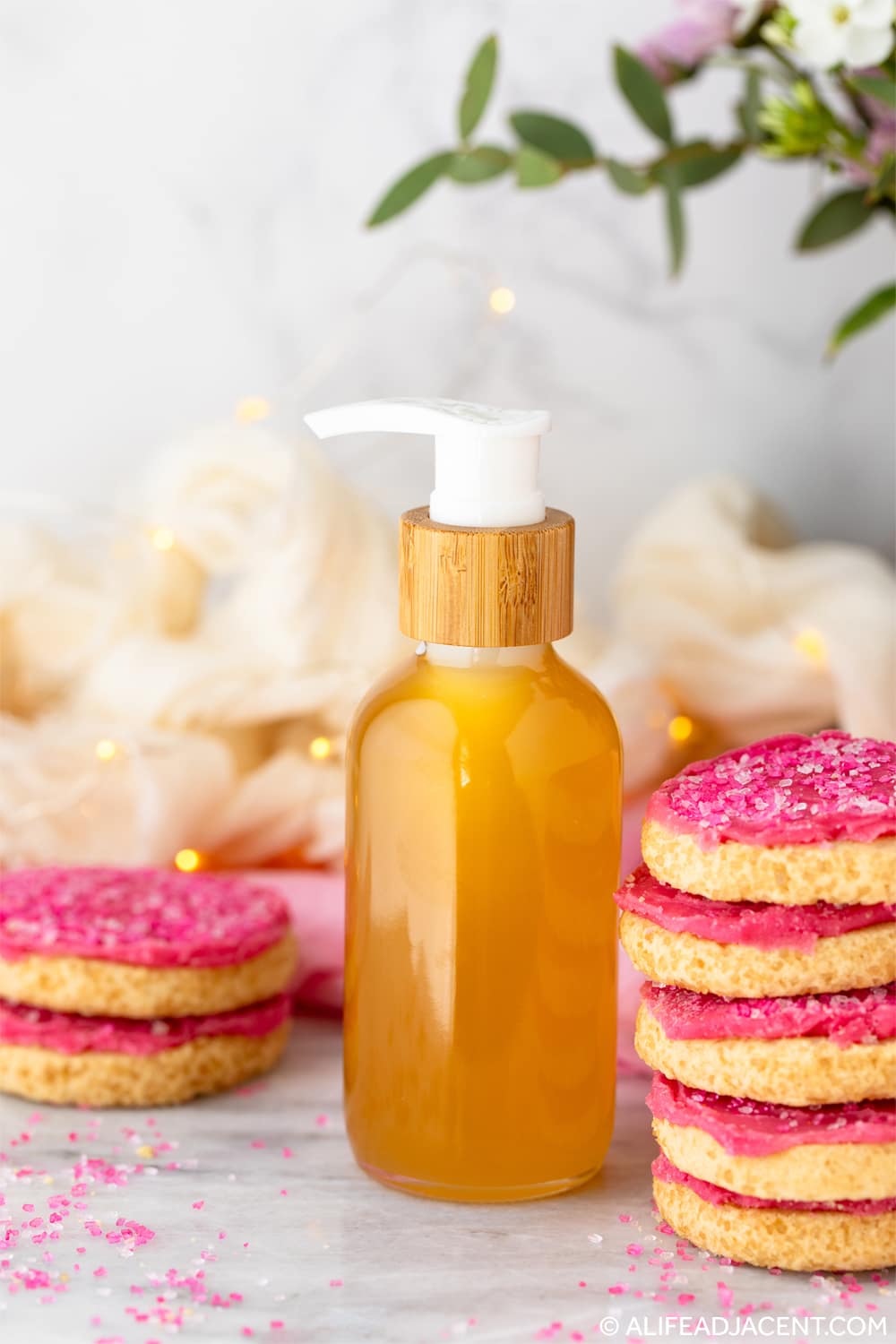 DIY sugar cookie perfumed body oil beside stacked frosted sugar cookies and soft fairy lights.