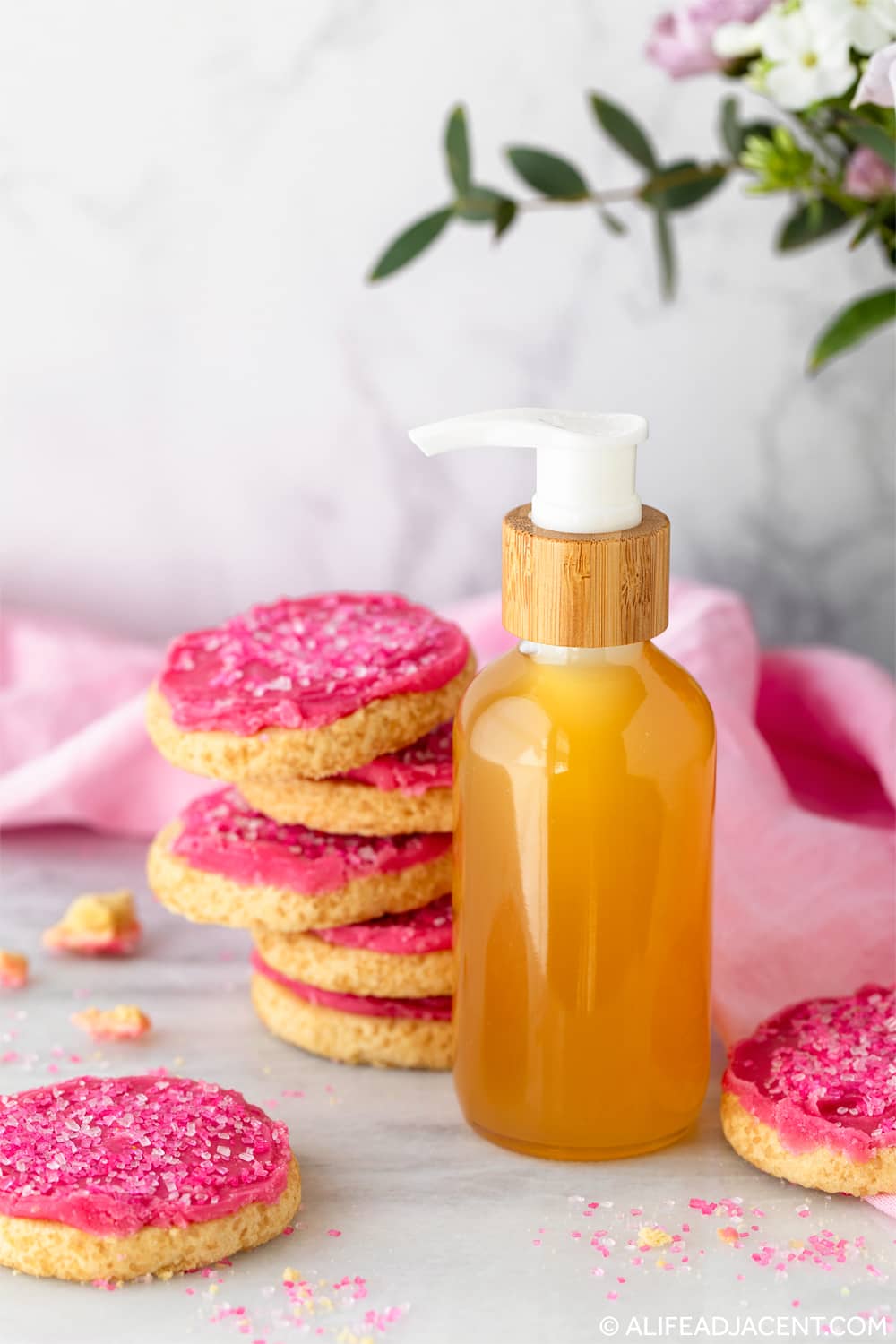 Homemade fragranced body oil displayed with pink frosted sugar cookies in a styled scene.