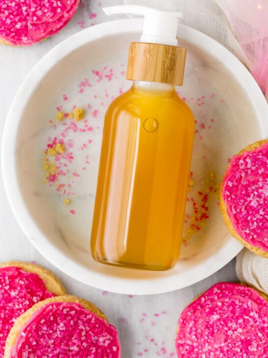 Sugar cookie body oil on marble plate surrounded by pink frosted sugar cookies.