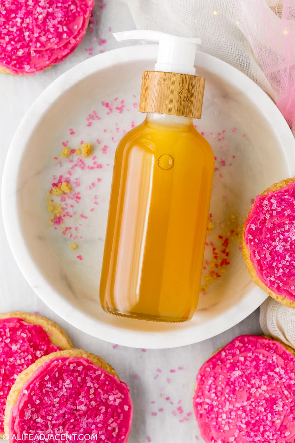 Sugar cookie body oil on marble plate surrounded by pink frosted sugar cookies.