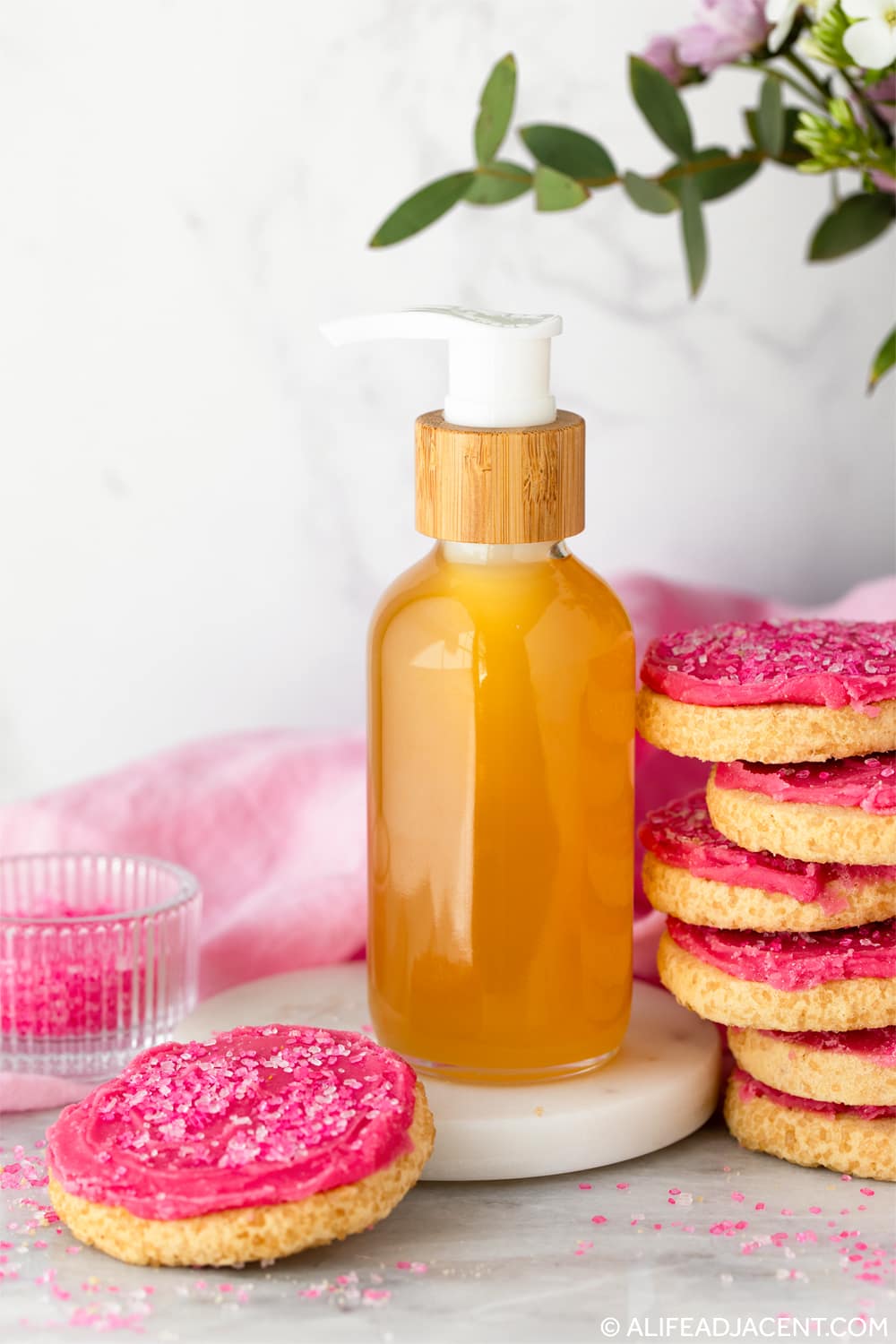 Sugar cookie-scented body oil in a glass pump bottle with stacked pink frosted sugar cookies.