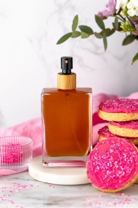 DIY sugar cookie body spray made with essential oils and natural perfumery oils, pictured beside a stack of pink frosted sugar cookies.