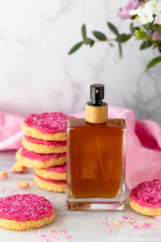 Homemade body spray with a natural sugar cookie scent, pictured with stacked pink iced cookies and pink fabric.