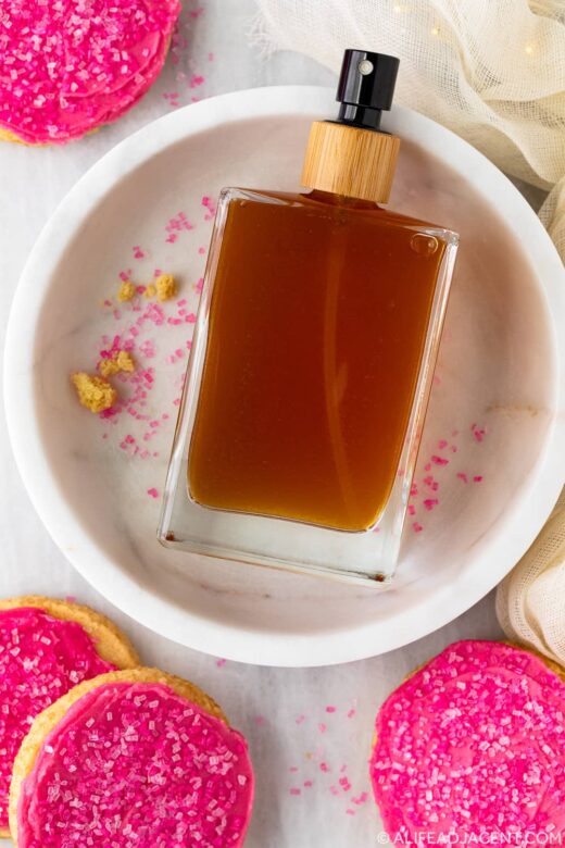 Sugar cookie body mist in a clear glass perfume bottle surrounded by pink frosted sugar cookies, photographed from above.